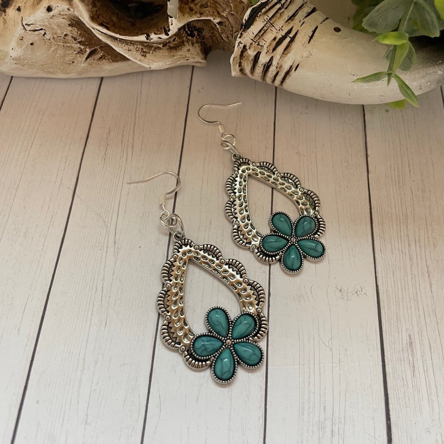 Turquoise flower earrings on a wooden surface with a skull and plant in the background