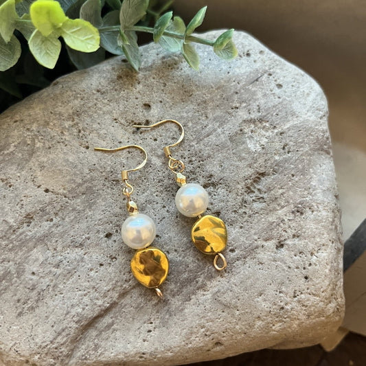 A pair of gold-tone dangle earrings with a white pearl and a faceted yellow bead, displayed on a textured gray stone with green leaves in the background.