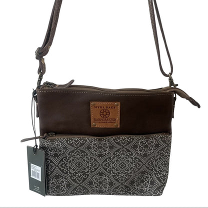 A women's crossbody bag by Stella Beach Manchester, featuring a dark brown leather top section with a tan brand patch, and a black and gray patterned fabric bottom section. It has a single shoulder strap and a hanging product tag.