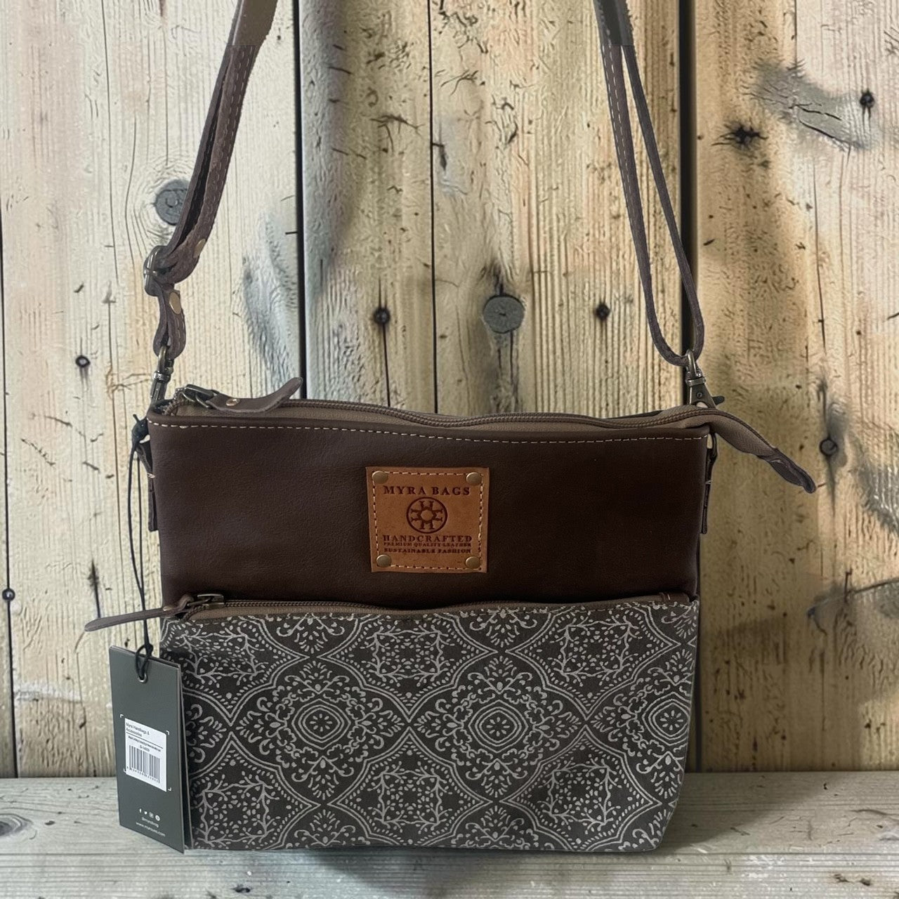 A Manchester crossbody bag with a dark brown leather top section and a black-and-white patterned lower pouch, displayed against a rustic wooden plank background.