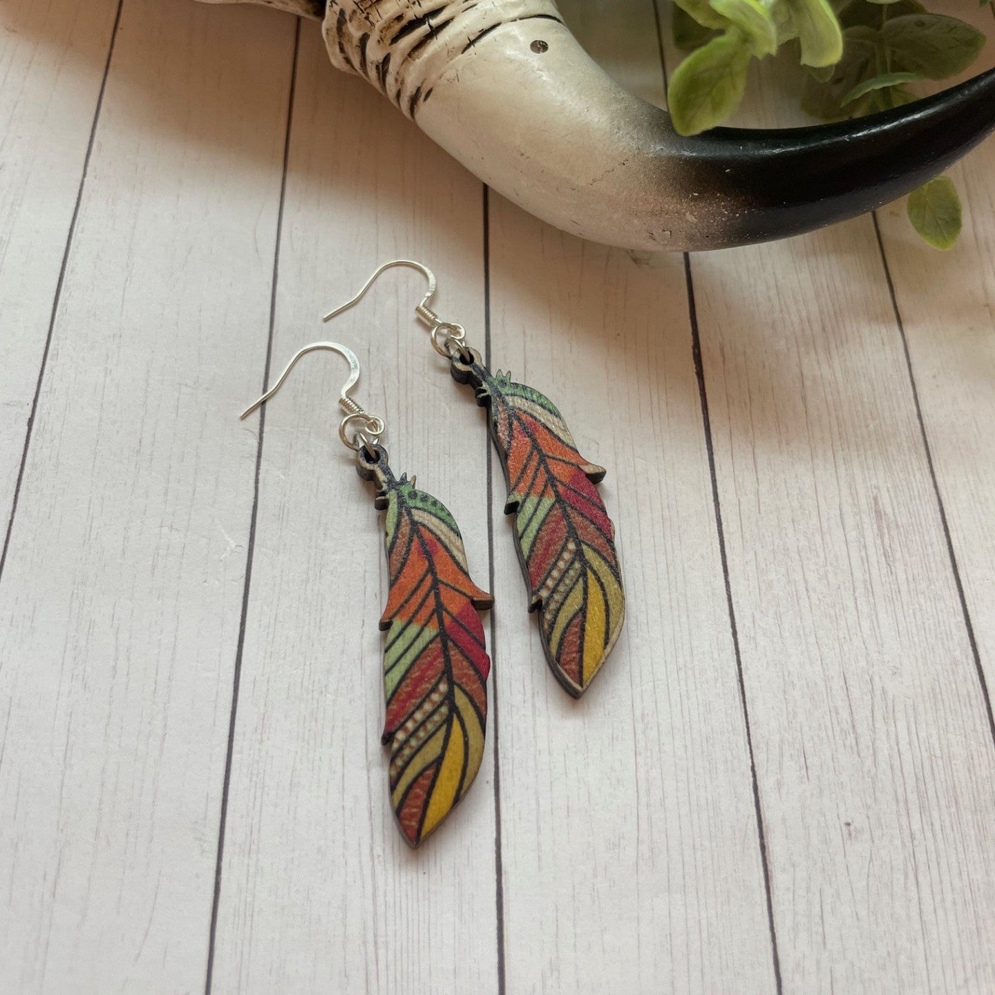 Colorful feather-shaped earrings on a wooden surface with a skull and plant in the background.