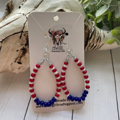 red white and blue teardrop shaped earrings in a teardrop shape on a white card with white wood background and a skull and greenery