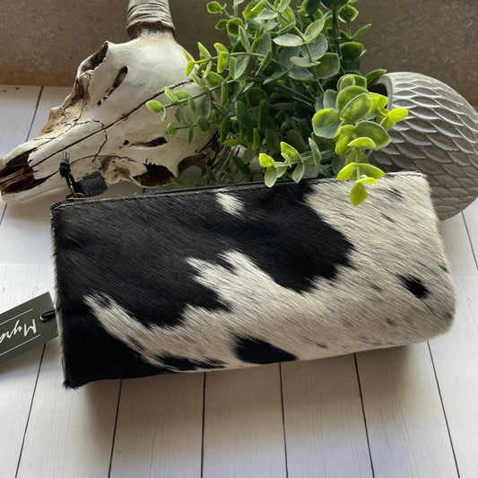 Black and white cowhide clutch bag with a plant and skull decoration on a light wooden floor.