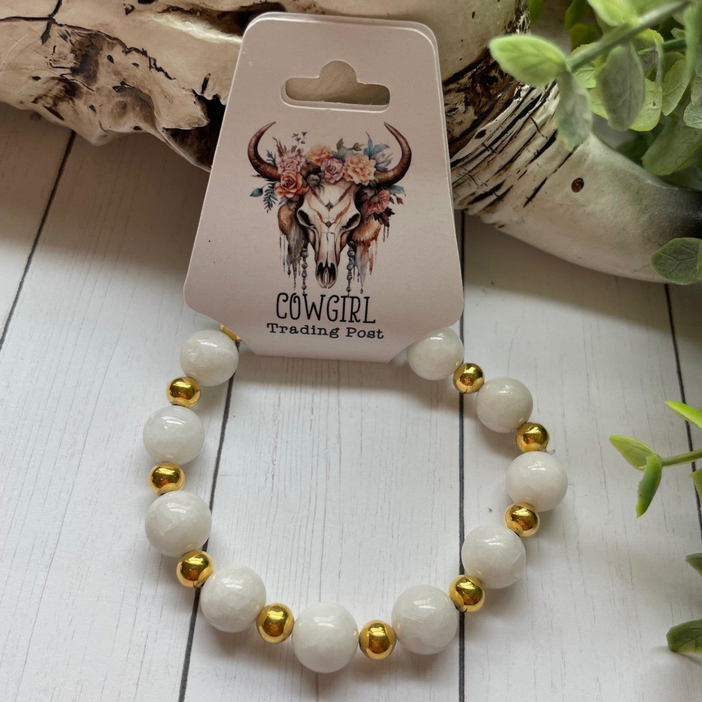 White beaded bracelet with gold accents on a white wooden surface, branded 'The Cowgirl Trading Post'.