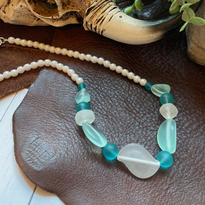 A beaded necklace with white pearl-like beads, light blue translucent stones, and teal-blue faceted beads, displayed on a brown leather surface with a skull and green plant in the background.
