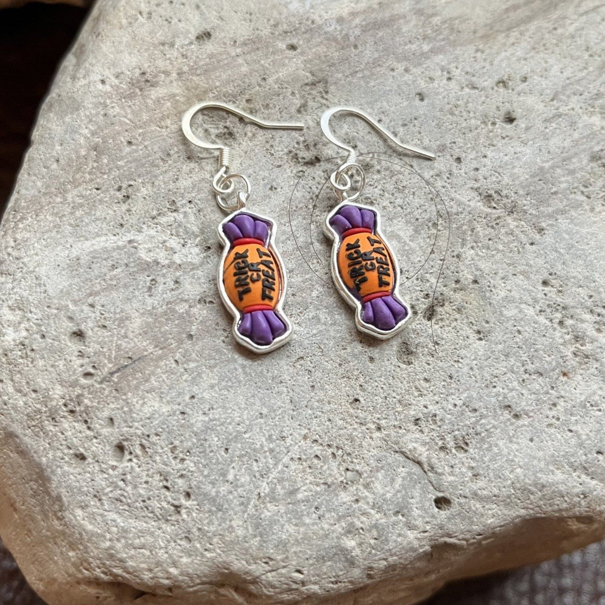 A pair of dangling earrings shaped like wrapped candies, with purple ends and orange centers featuring black Japanese characters, displayed on a textured gray stone.