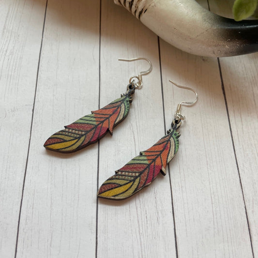 Colorful feather-shaped earrings on a wooden surface with a decorative elephant and plant in the background.
