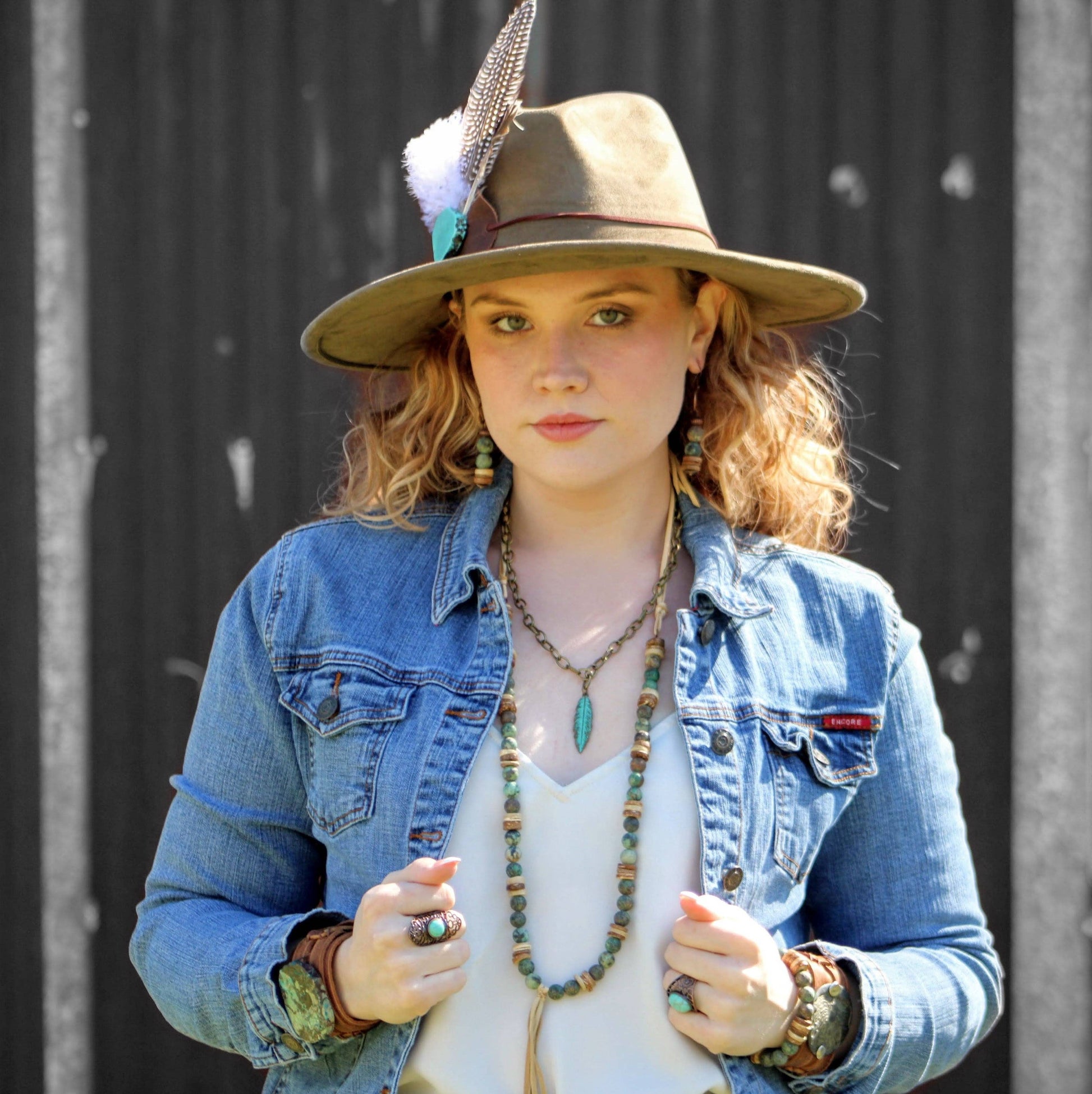 Woman wearing a denim jacket, white top, and hat with a feather against a black background