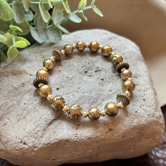 A beaded stretch bracelet with metallic gold and bronze beads, displayed on a textured gray stone with green leaves in the background.