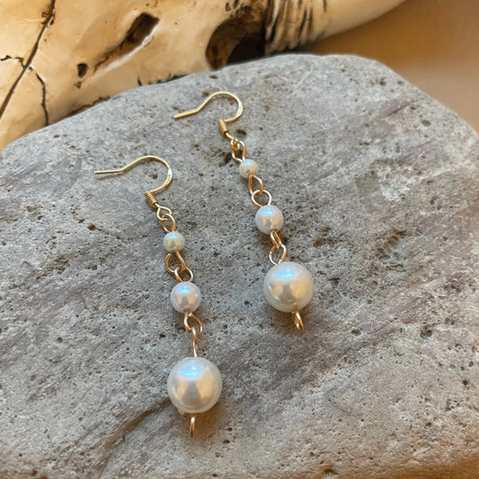 round pearl beads and gold wire earrings dangling from a gold ear wire, on a beige colored stone with a cow or bull skull in the background,.