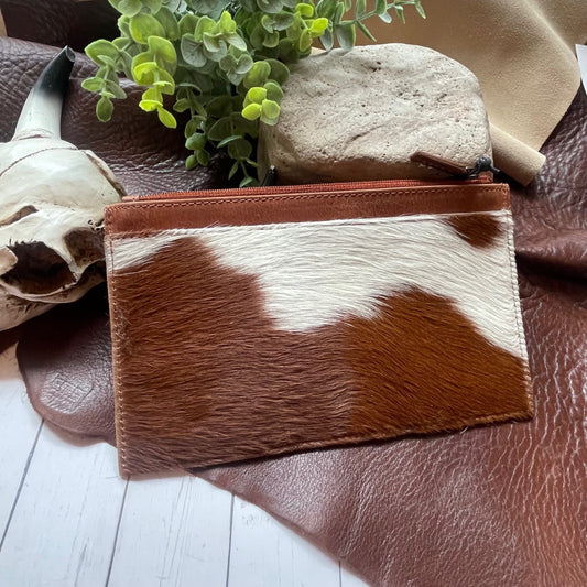A small zip pouch made from brown and white cowhide hair, styled on a brown leather surface with a skull, green leaves, a stone, and a beige fabric background.
