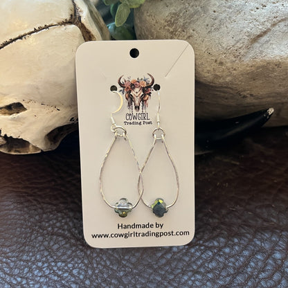 A pair of silver teardrop dangle earrings with small iridescent beads, displayed on a cream-colored Cowgirl Trading Post card, set against a rustic background of a skull, stone, and dark brown leather.