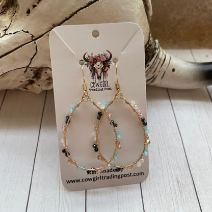 Pair of gold earrings with beads on a Cowgirl Trading Post card, placed on a wooden surface.
