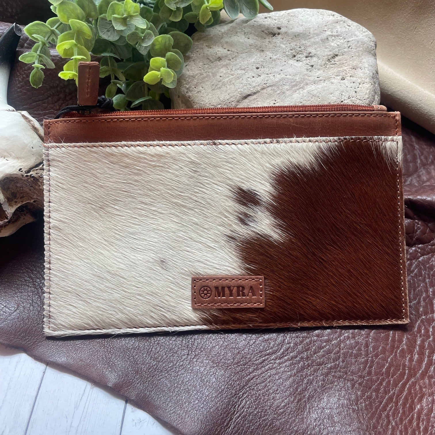 A zippered pouch with a brown and cream cowhide pattern, featuring a brown leather brand patch that reads 'MYRA' and a matching brown leather zipper trim, displayed on a brown leather surface with decorative stones and a small green plant.