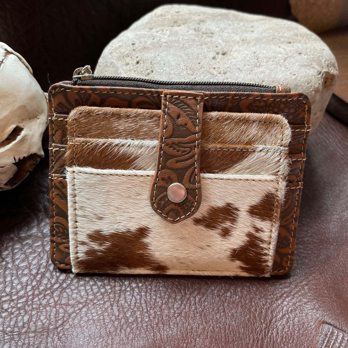 A small bifold wallet made from brown embossed leather and cowhide, featuring a front card pocket with a snap tab closure, photographed on a brown leather surface with decorative props.
