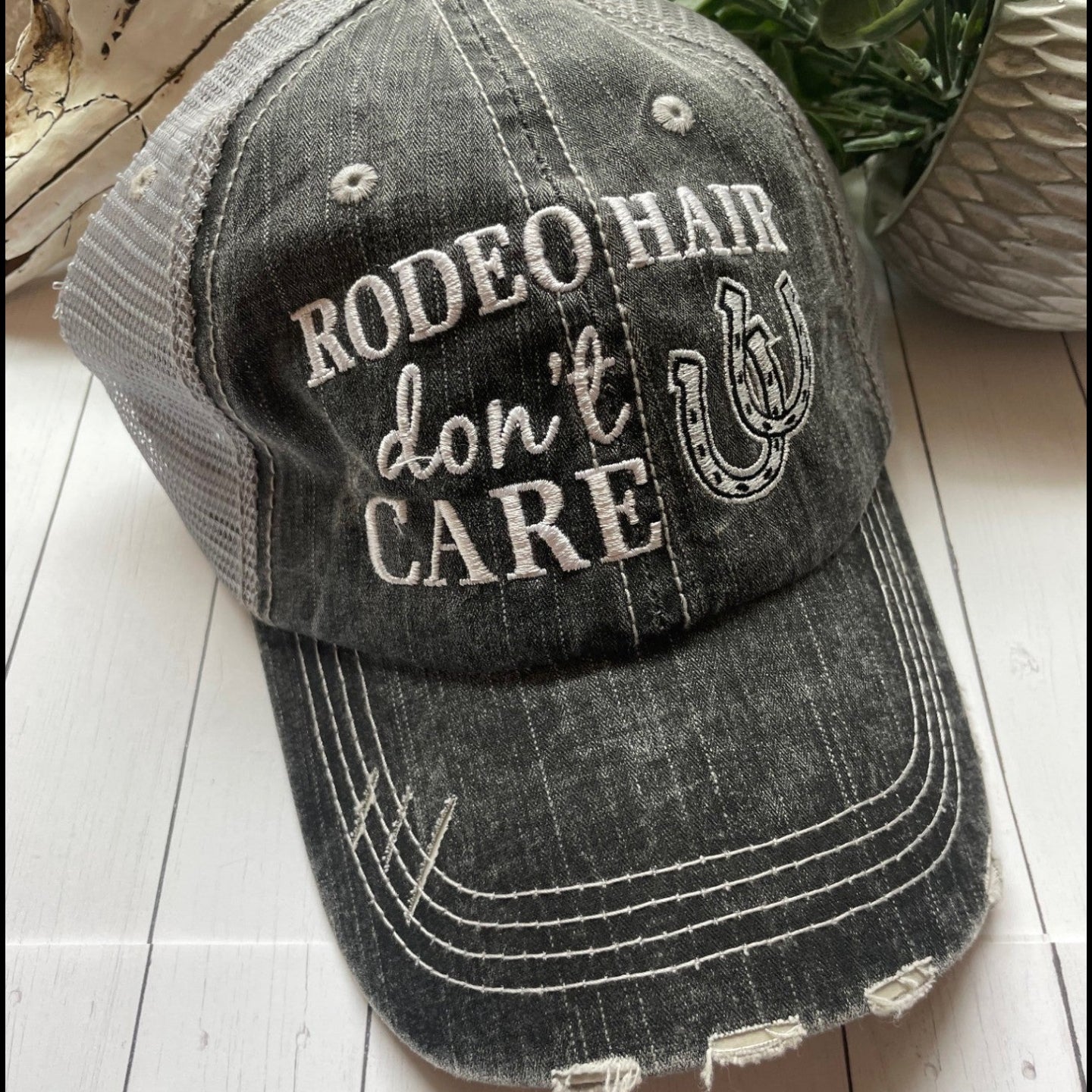 Black distressed baseball cap with 'Rodeo Hair Don't Care' text on a wooden surface.