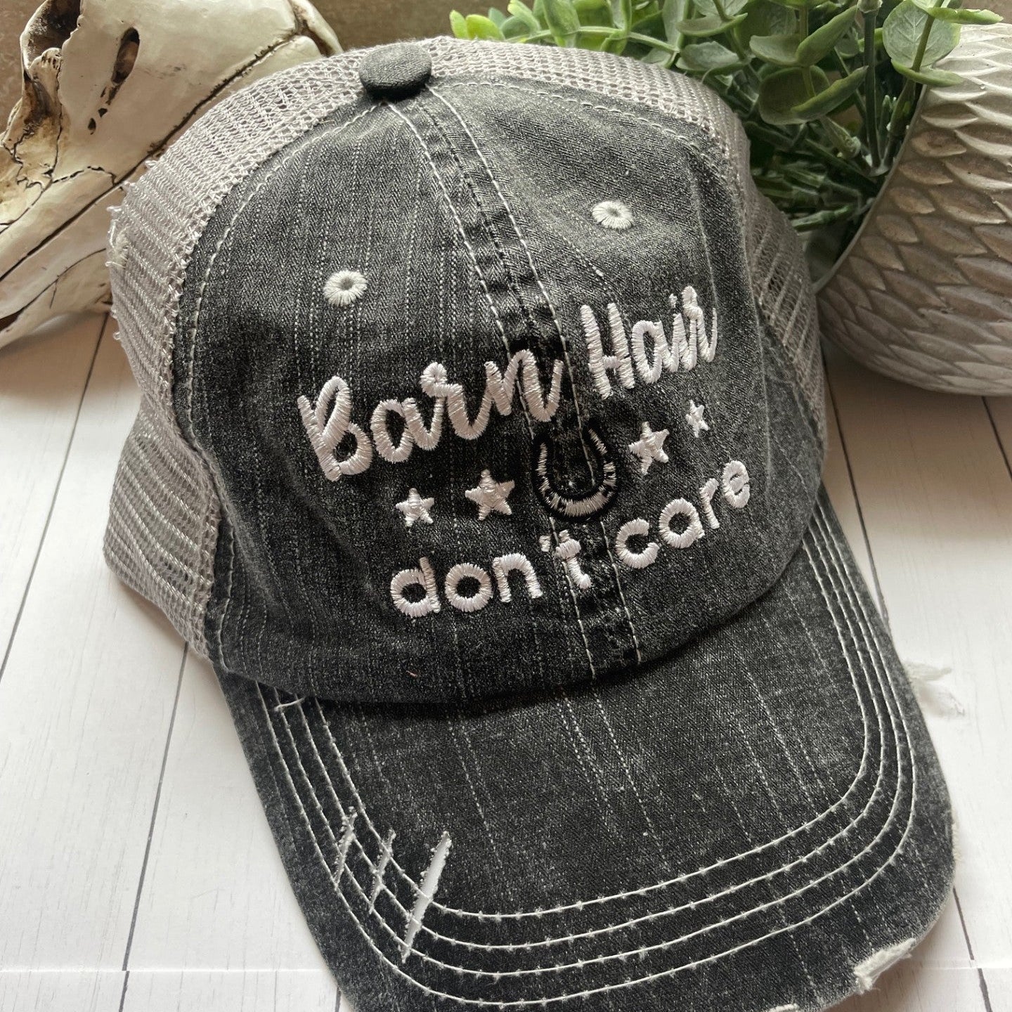 Black distressed cap with 'Barn Hair don't care' text on a wooden surface with plants in the background