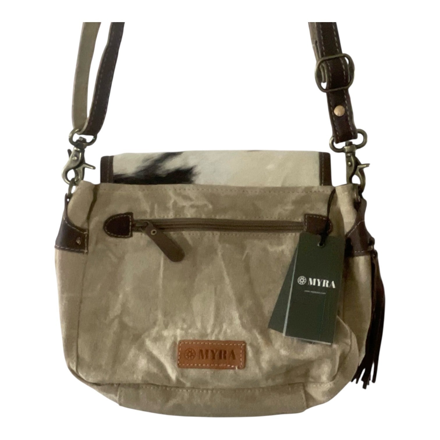 A MYRA brand shoulder bag in beige canvas with brown leather trim, a black and white cow print accent panel, and a green hangtag, shown against a plain white background.