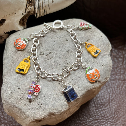 A silver charm bracelet with Halloween-themed enamel charms, displayed on a gray stone with a dark brown textured surface and a bone prop in the background.