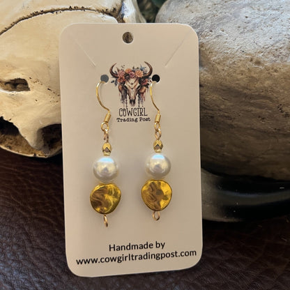 Cowgirl Trading Post handmade dangle earrings with gold-toned metal, white pearl beads, and textured gold disc charms, displayed on a branded white card against a rustic background of a skull, stone, and greenery.