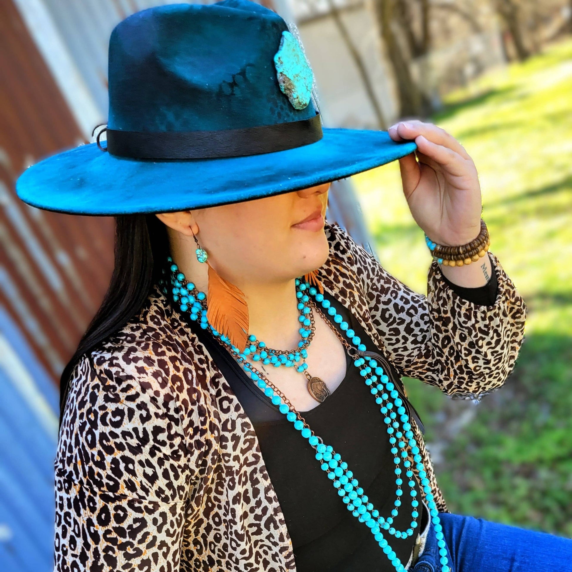 Person wearing a blue hat with a floral detail, leopard print jacket, and turquoise jewelry outdoors.