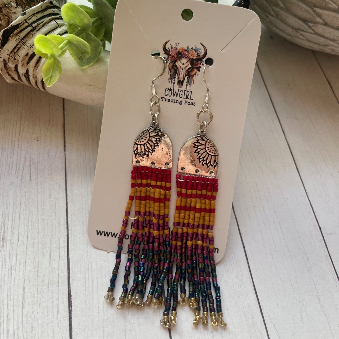 Pair of beaded earrings on a branded card with a wooden surface and plant in the background