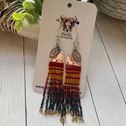 Pair of beaded earrings on a branded card with a wooden surface and plant in the background