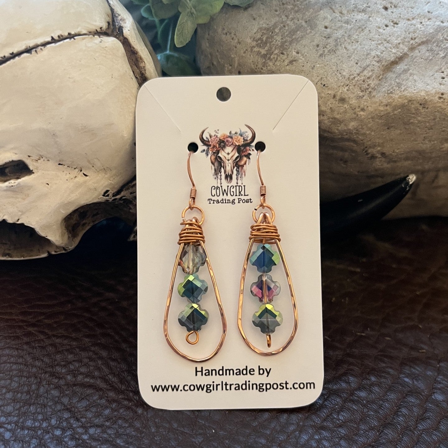 A pair of handmade teardrop dangle earrings with copper wire frames and iridescent green, blue, and purple faceted beads, displayed on a white cowgirl trading post card. The card is set against a rustic background featuring a skull, green leaves, and a dark textured surface.