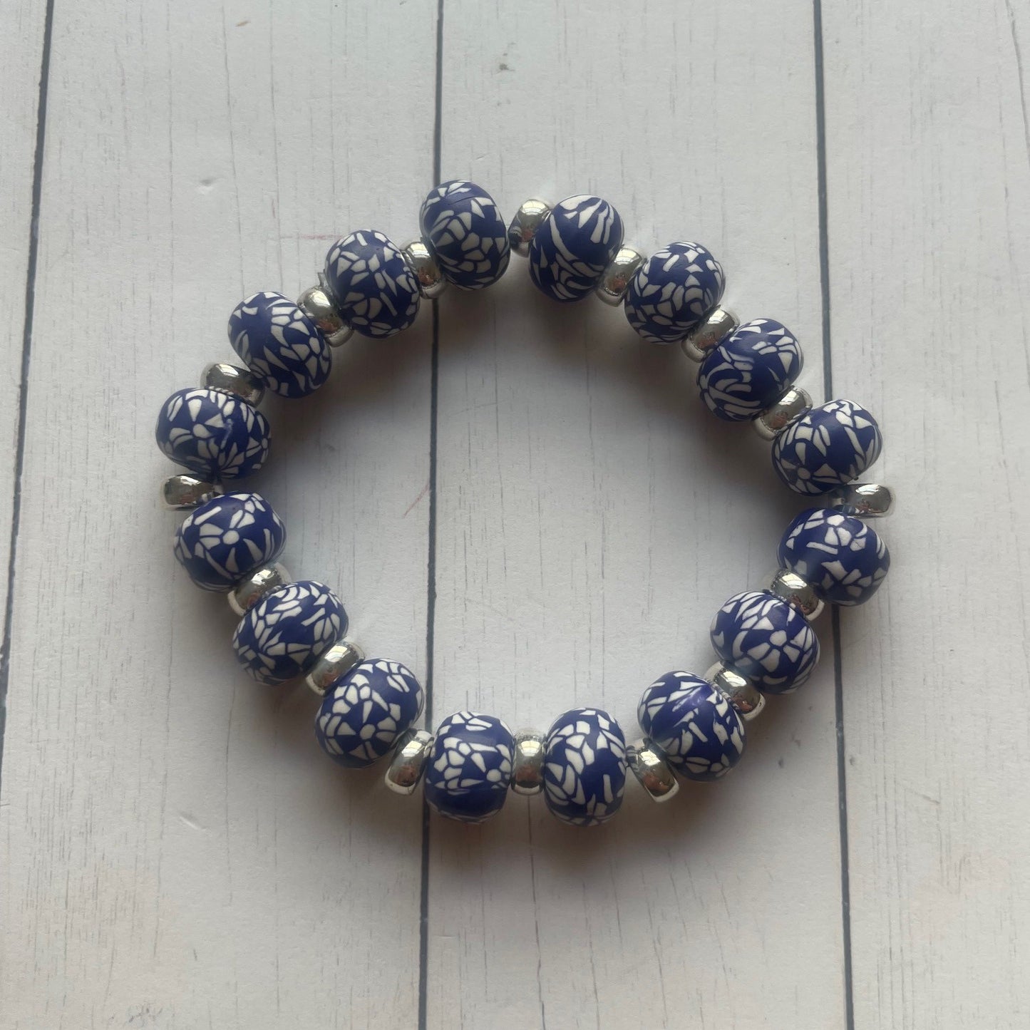 A beaded stretch bracelet with glossy blue and white patterned round beads separated by small silver spacer beads, displayed on a white wood plank surface.