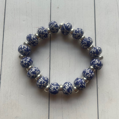 A beaded stretch bracelet with glossy blue and white patterned round beads separated by small silver spacer beads, displayed on a white wood plank surface.