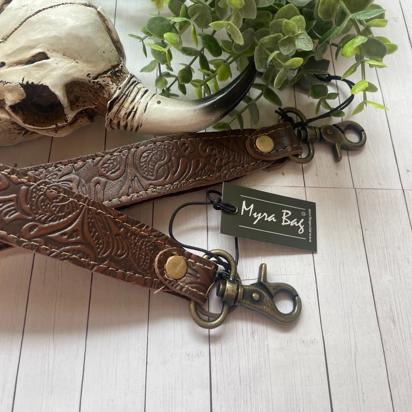 Brown leather strap with gold hardware on a white wooden surface, accompanied by a plant and a tag labeled 'Myra Bag'.