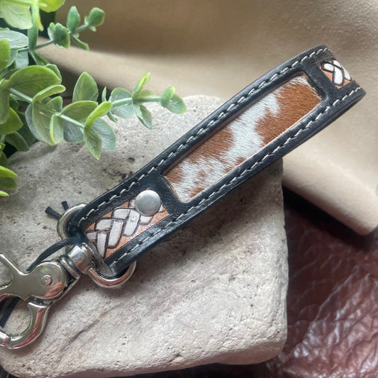 A leather strap accessory with a brown and white cowhide pattern center, black leather trim, silver-toned metal hardware, and decorative woven leather details, displayed on a stone block with green leaves and a tan fabric background.