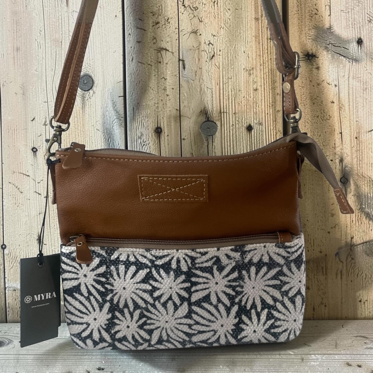 A brown leather and patterned fabric shoulder bag with a black hangtag showing the brand MYRA, set against a rustic wooden plank background.
