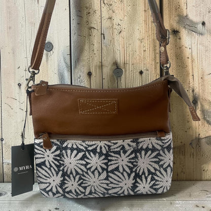A brown leather and patterned fabric shoulder bag with a black hangtag showing the brand MYRA, set against a rustic wooden plank background.