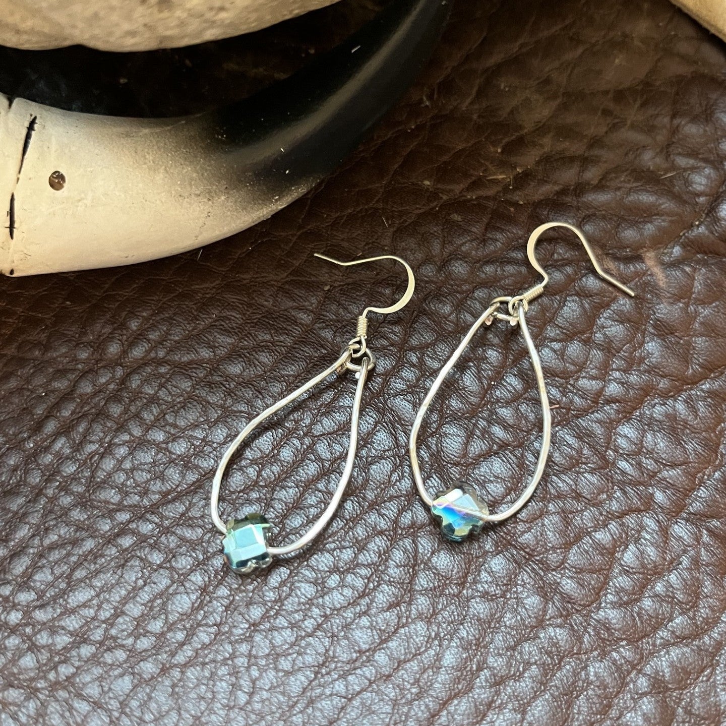 A pair of silver teardrop dangle earrings with small light blue and white inlaid bead accents, displayed on a brown textured leather surface next to a black and white horn.