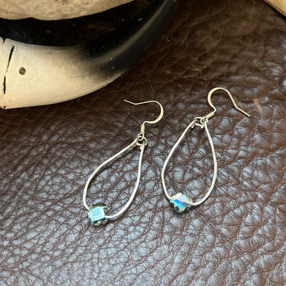 A pair of silver teardrop dangle earrings with small light blue and white inlaid bead accents, displayed on a brown textured leather surface next to a black and white horn.