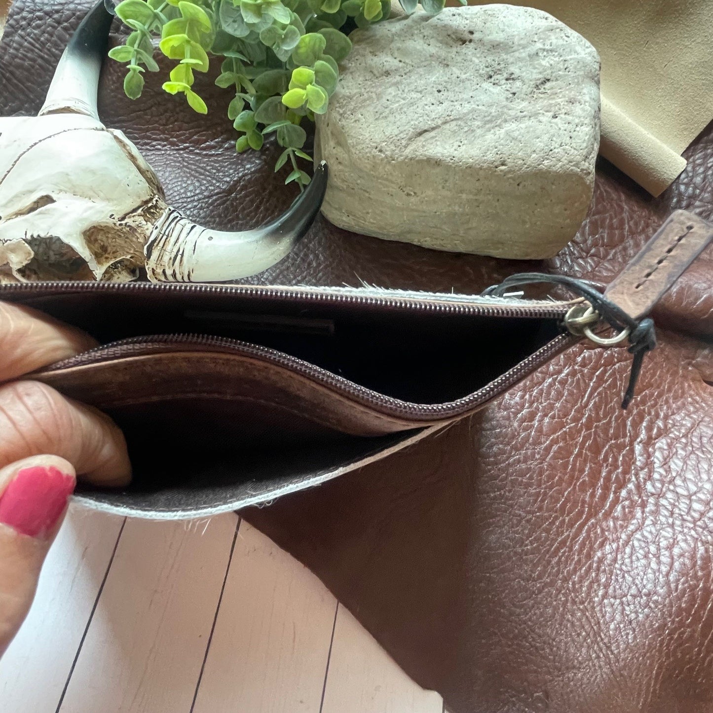 A close-up of a brown leather zip pouch being held open by a hand with pink nail polish, set against a brown leather background with decorative props including a small plant, a stone, and a horn.