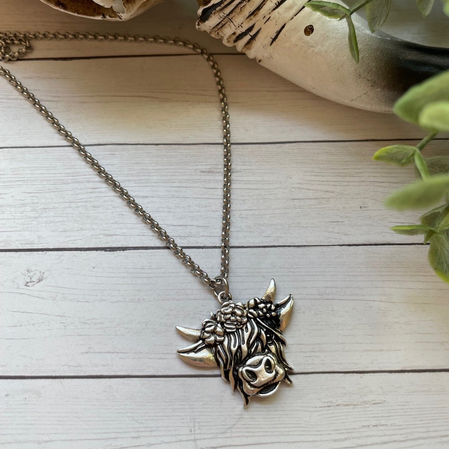 Silver necklace with a bull highland cow head pendant on a wooden surface with a plant in the background