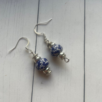 A pair of handmade dangle earrings featuring blue and white patterned glass beads with silver-toned metal hooks, displayed on a white wooden surface.