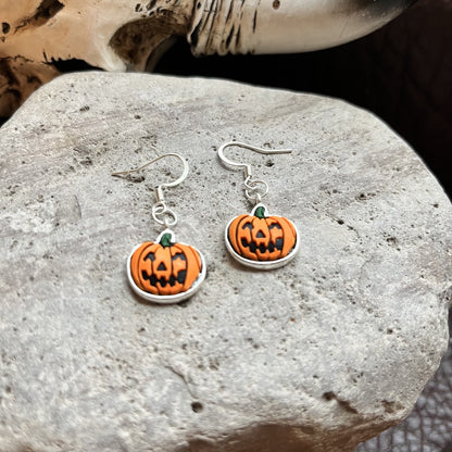 A pair of Halloween-themed drop earrings featuring orange pumpkin charms with black carved facial features and a small green stem, suspended from silver fishhook wires. The earrings are displayed on a textured gray stone surface.