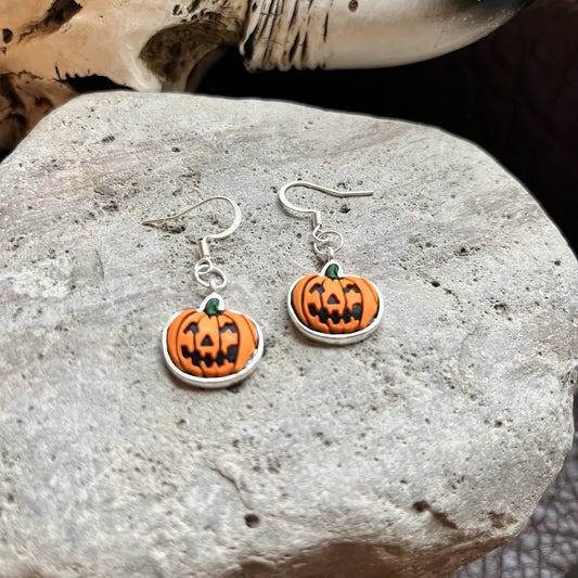 A pair of Halloween-themed drop earrings featuring orange pumpkin charms with black carved facial features and a small green stem, suspended from silver fishhook wires. The earrings are displayed on a textured gray stone surface.