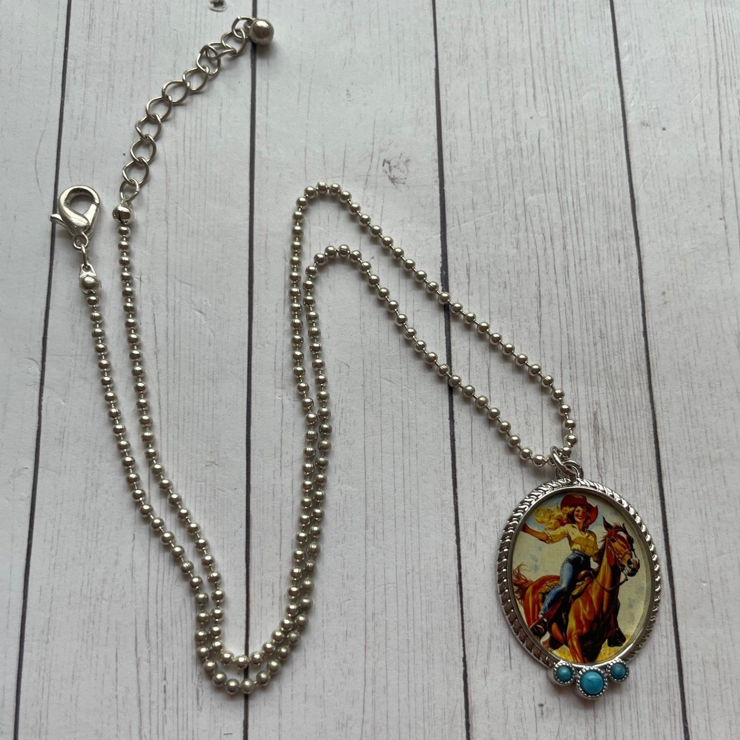 A silver-toned beaded chain necklace with an oval pendant featuring a colorful western-style illustration of a rider on a horse, accented with small turquoise-like beads at the bottom, displayed on a white wood plank background.
