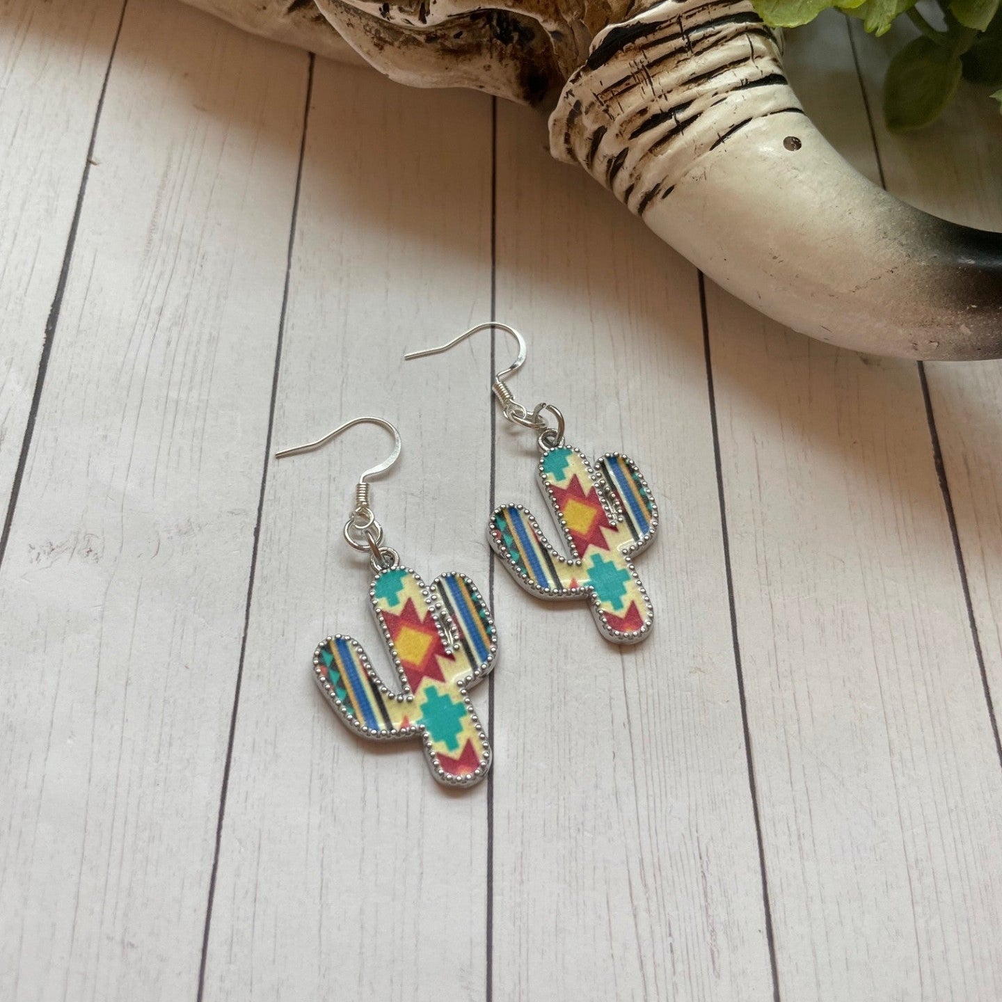 Colorful cactus-shaped earrings on a wooden surface with a decorative stone and plant in the background.