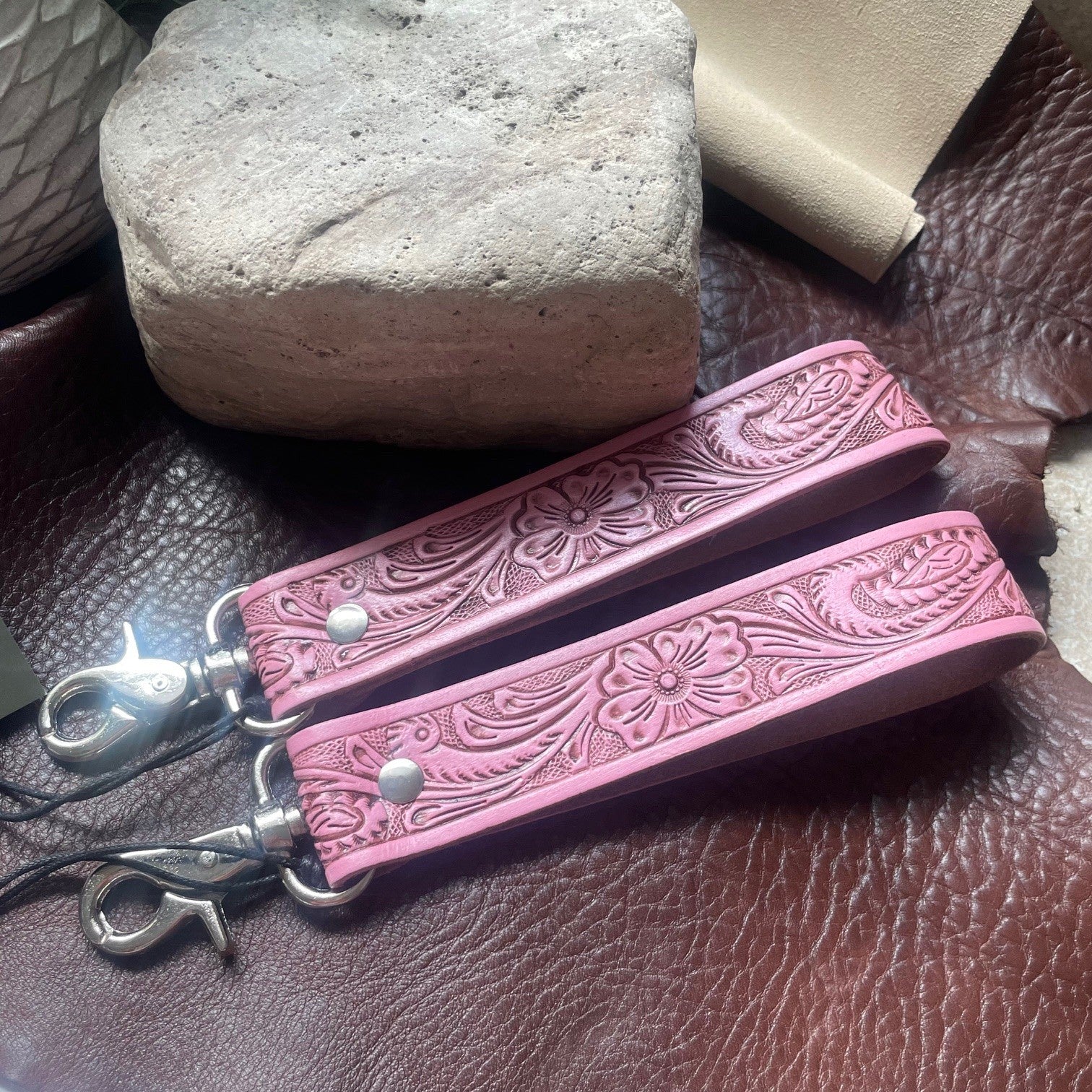 Two pink tooled leather strap keychains with silver-tone hardware, displayed on a brown textured leather surface with a stone and beige material in the background.