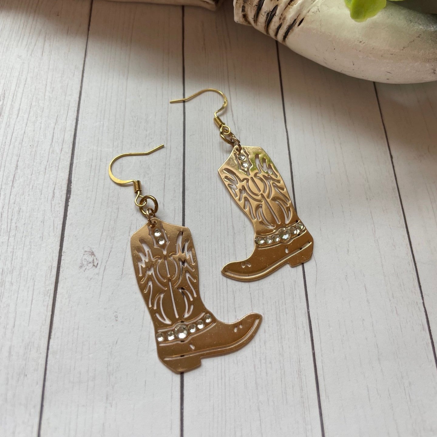 Cowboy boot earrings on a wooden surface with a decorative skull and greenery in the background.