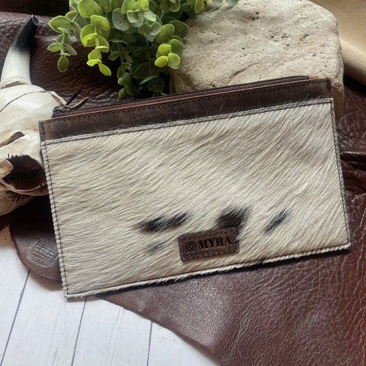 A western-style zippered pouch featuring a cream and black cowhide patterned front with dark brown leather trim and a 'MYRA' brand patch, displayed on a brown leather surface with a plant in the background.