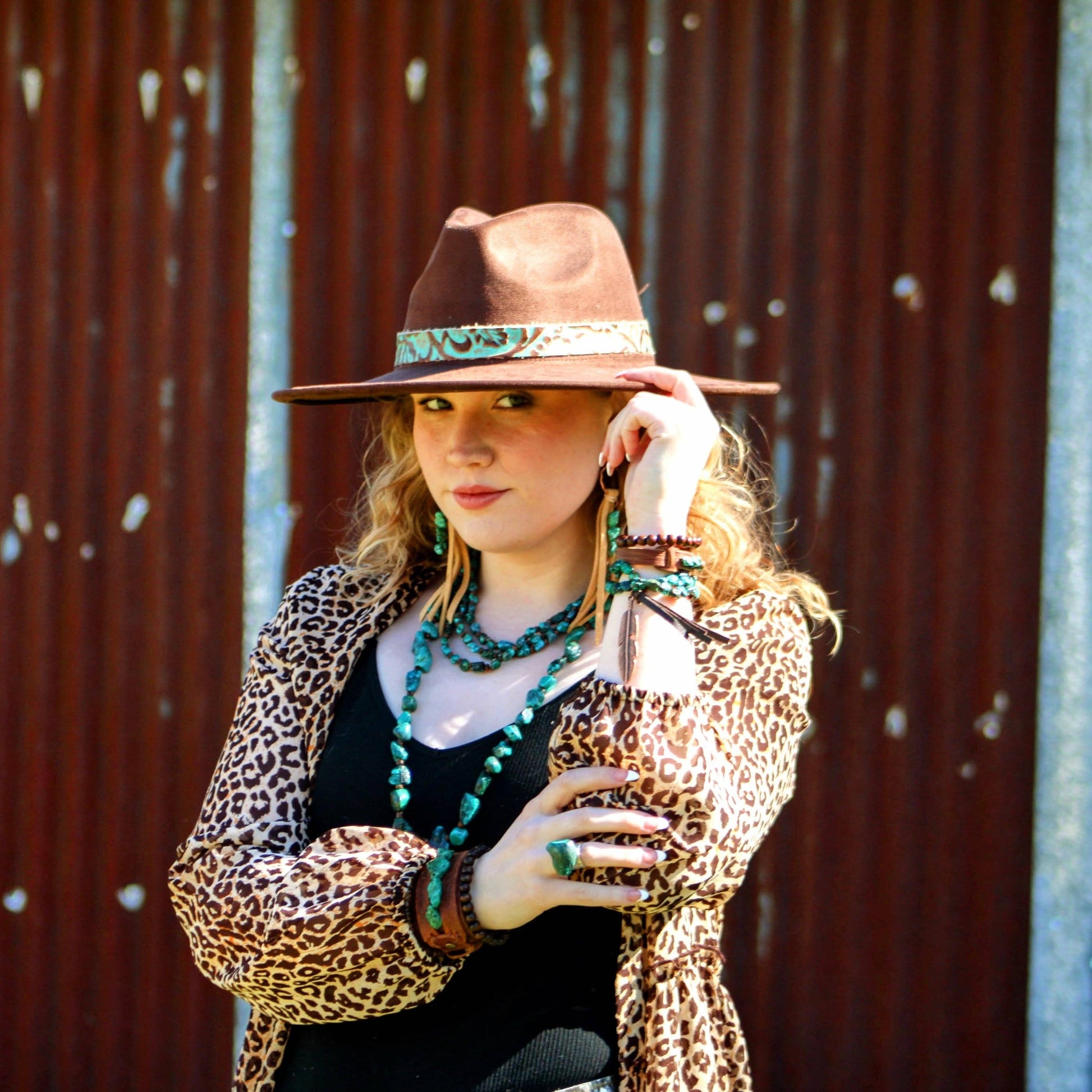 Woman wearing a brown hat with a band, leopard print jacket, and multiple necklaces against a corrugated metal background.