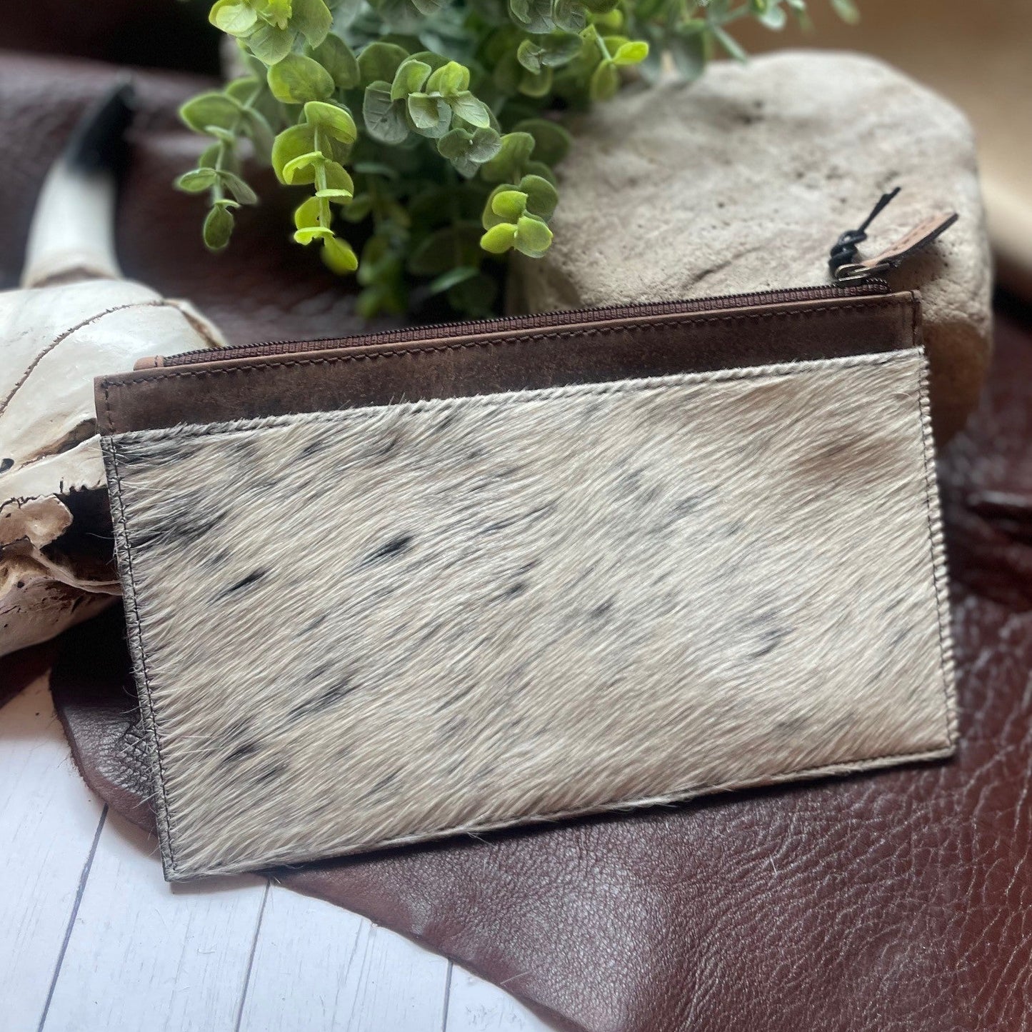 A small zip-around pouch or card holder with a natural cowhide-textured front panel in off-white and tan with dark speckles, plus a dark brown trim and zipper, styled on a brown leather surface with a plant and stones in the background.