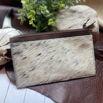 A small zip-around pouch or card holder with a natural cowhide-textured front panel in off-white and tan with dark speckles, plus a dark brown trim and zipper, styled on a brown leather surface with a plant and stones in the background.