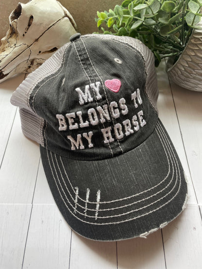 Black distressed cap with 'My Heart Belongs to My Horse' text on a wooden surface with a plant in the background.